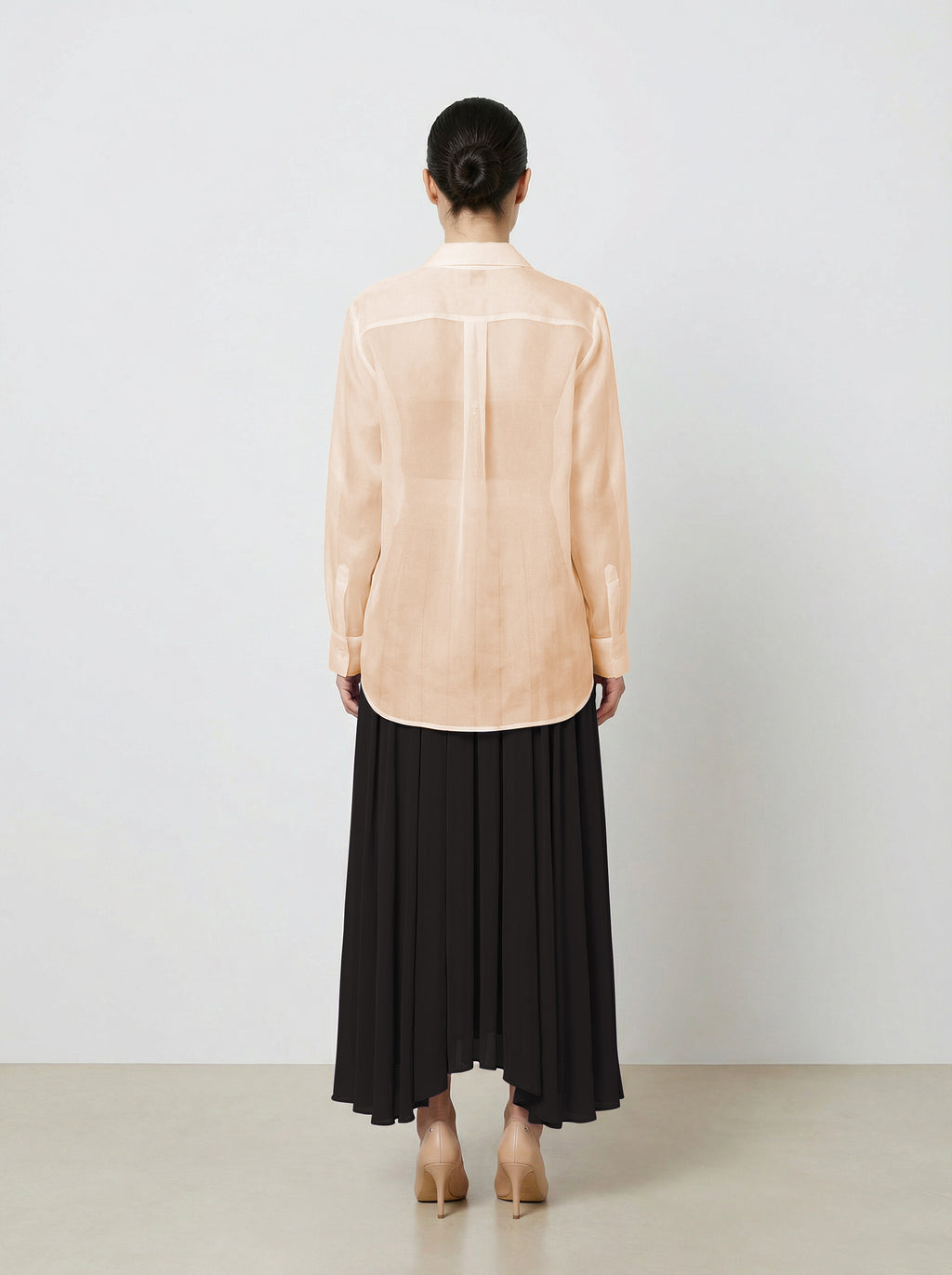 Woman wearing a light pink  organza blouse and black pleated skirt on a plain background back untucked view