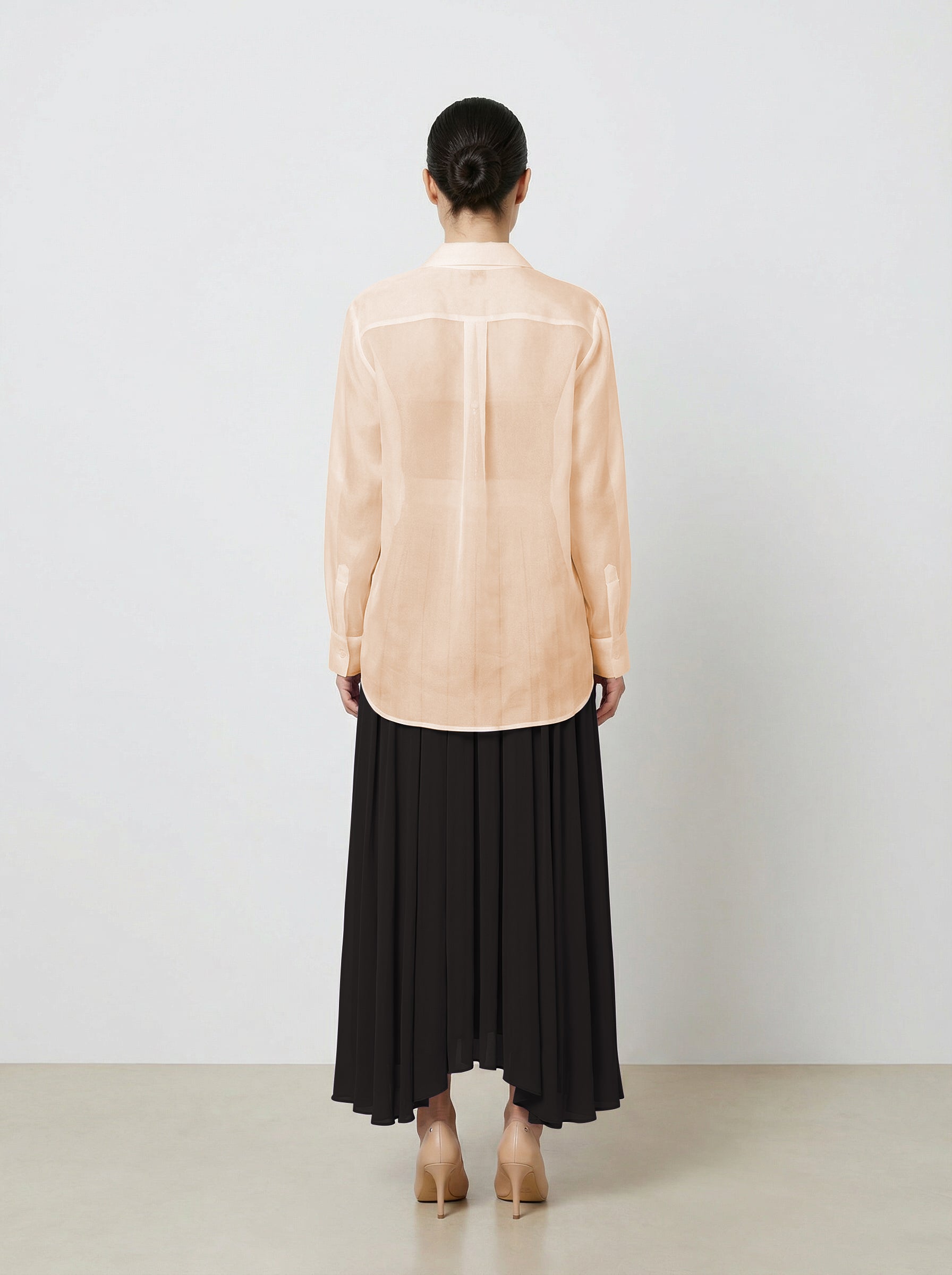 Woman wearing a light pink  organza blouse and black pleated skirt on a plain background back untucked view