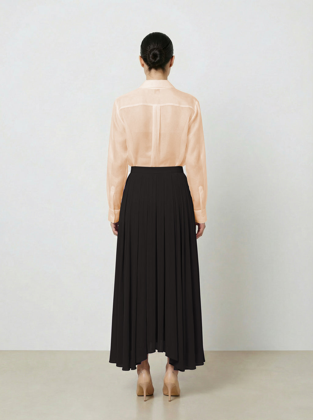 Woman wearing a light pink  organza blouse and black pleated skirt on a plain background back view