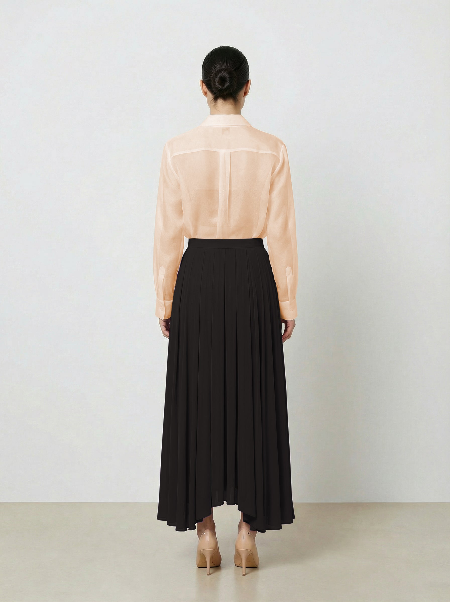 Woman wearing a light pink  organza blouse and black pleated skirt on a plain background back view