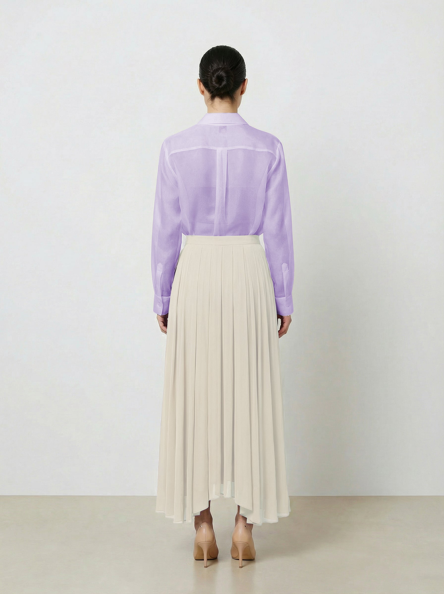 Woman wearing a light purple  translucent organza blouse and creme pleated skirt on a white background back view