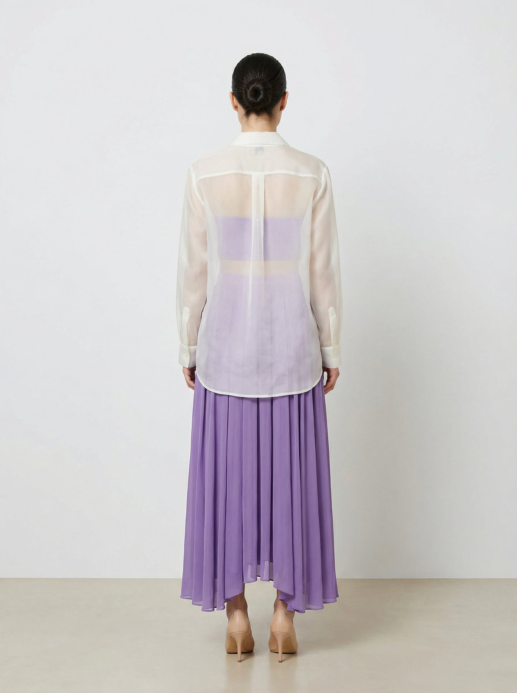Person wearing a sheer white top with a purple panel and a long purple skirt on a plain background