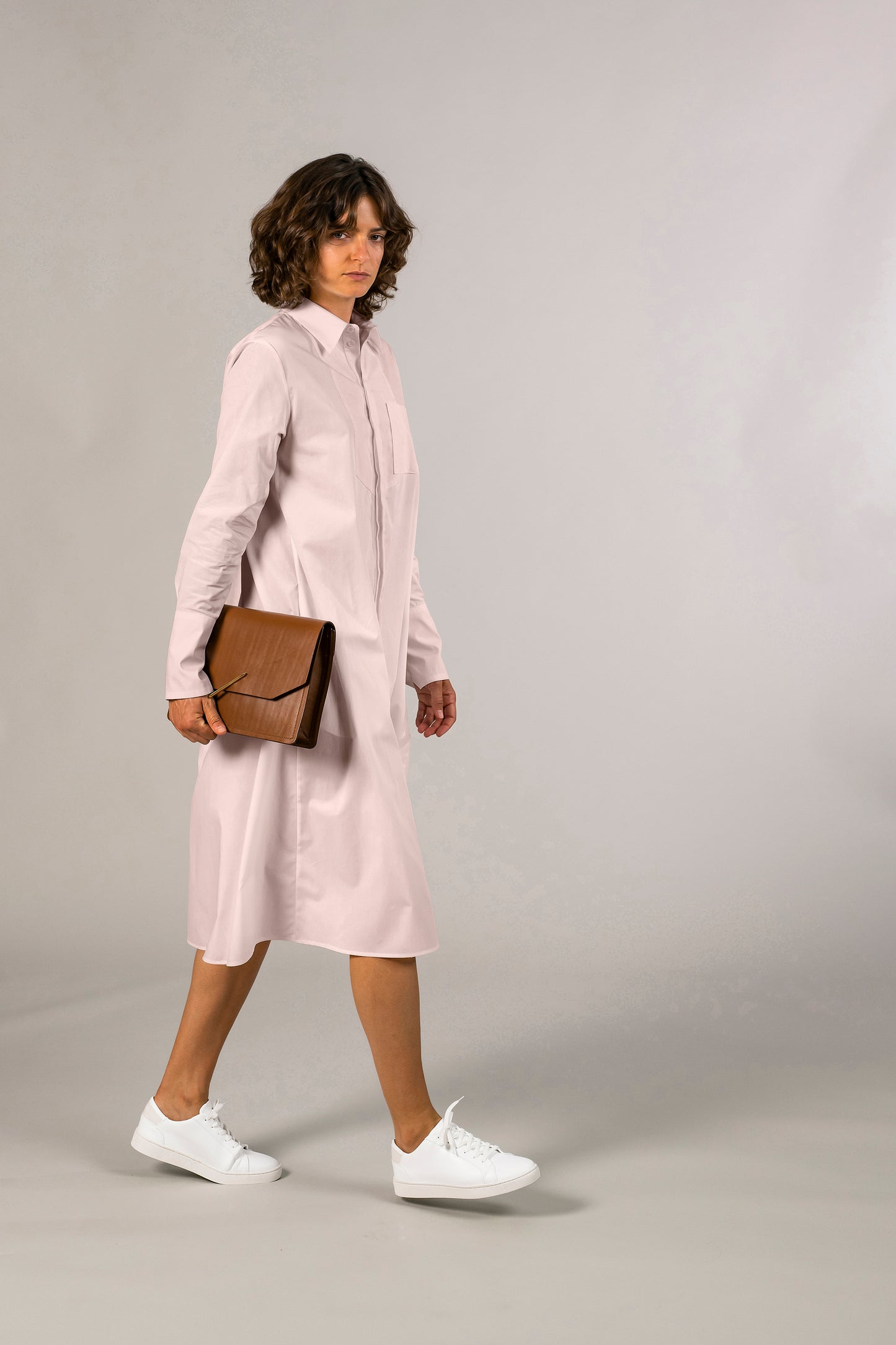 Side view of womens pink tuxedo shirt dress, 100% sustainable cotton poplin from Italy, long sleeve, button down knee length. Ensembled with Large brown leather clutch.